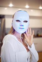 Portrait of a beautiful woman performing a light therapy facial mask procedure at home. photo