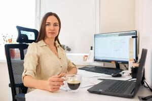 retrato de un remoto obrero. un hermosa mujer se sienta a un mesa en frente de un computadora y diseños un edificio. confidente joven dama persona de libre dedicación trabajando desde hogar. foto