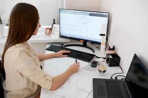 Portrait of a remote worker. A beautiful woman sits at a table in front of a computer and designs a building. Confident young lady freelancer working from home. photo