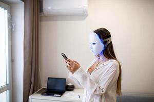 retrato de un hermosa mujer comunicado en el teléfono y ejecutando un ligero terapia facial máscara procedimiento a hogar. foto