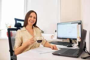 Portrait of a remote worker. A beautiful smiling brunette girl sits at a table in front of a computer and works with a layout. Confident young lady freelancer working from home. photo