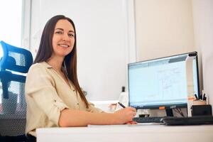 Portrait of a remote worker. A beautiful smiling brunette girl sits at a table in front of a computer and works with a layout. Confident young lady freelancer working from home. photo