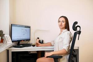 Portrait of a remote worker. A beautiful woman sits at a table in front of a computer and designs a building. Confident young lady freelancer working from home. photo