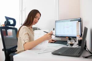 Portrait of a remote worker. An adult woman sits at a table in front of a computer and draws up a costly estimate. Confident young lady freelancer working from home. photo