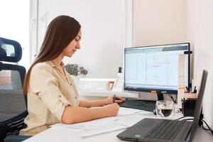 Portrait of a remote worker. A beautiful woman sits at a table in front of a computer and designs a building. Confident young lady freelancer working from home. photo