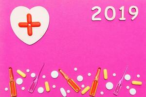 Pharmacy concept Happy New Year 2019 for medical and health concept. pharmaceutical background. Pills, ampoules and a wooden heart with a red cross from tablets on a pink background. photo