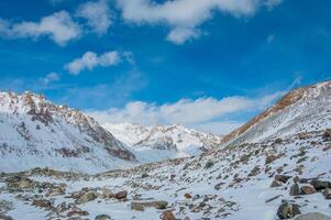 Snow-covered peaks rise against blue sky, rocky slopes meet a valley floor, scattered stones dot the white expanse. photo