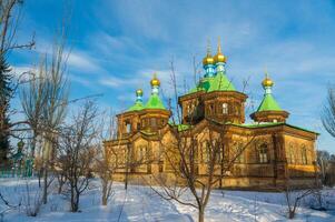 Ornate Holy Trinity wooden Church stands in Karakol, golden domes gleaming and and crosses. Leafless trees and snow foreground against a clear blue sky, showcasing winter in a historic setting photo