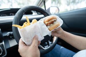 Asian lady holding hamburger and French fries to eat in car, dangerous and risk an accident. photo