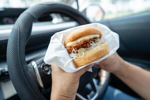 Asian lady holding hamburger to eat in car, dangerous and risk an accident. photo