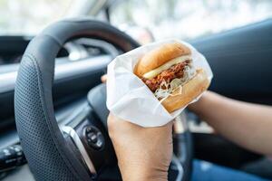 asiático dama participación hamburguesa a comer en auto, peligroso y riesgo un accidente. foto