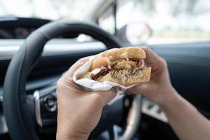 asiático dama participación hamburguesa y francés papas fritas a comer en auto, peligroso y riesgo un accidente. foto