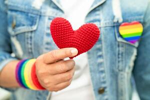 Asian lady wearing rainbow flag wristbands and hold red heart, symbol of LGBT pride month celebrate annual in June social of gay, lesbian, bisexual, transgender, human rights. photo