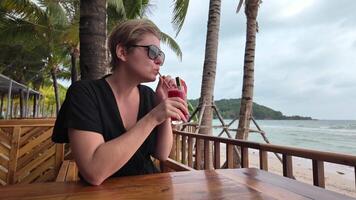 een ontspannen vrouw genieten van een verfrissend watermeloen drinken Bij een tropisch strand bar, met palm bomen en oceaan golven in de achtergrond, beeltenis vakantie vrije tijd video