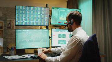 Businessman in home office drinking coffee and talking on headphones about financial crisis. Four big displays with different stock market data video