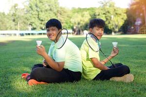 An Asian boy holds a badminton racket and a white shuttlecock while playing badminton with friends on the park lawn in the evening after returning from school. photo