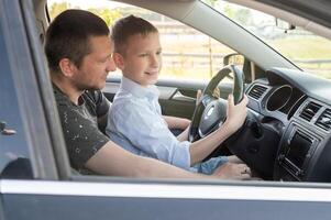 papá y hijo en el frente asiento de el coche a el rueda. el chico sostiene el direccion rueda foto