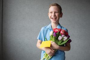 un linda chico sonrisas y soportes con un regalo y tulipanes dar un ramo de flores de flores foto