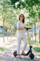 Joyful adorable senior woman using a scooter while riding in the park. Modern woman, a new generation. Healthy cheerful senior retired lady. Concept of Age Inclusivity and ecological transport photo