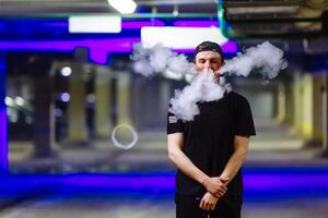 Man in cap smoke an electronic cigarette and releases clouds of vapor performing various kind of vaping tricks photo