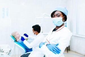 Young African american female dentist in mask in front of dentist checking teeth to patient at clinic. Dental clinic concept. photo