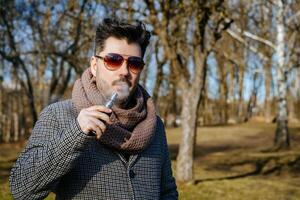 Confident young bearded male in sunglasses vape electronic cigarette. A young handsome bearded hipster man vaping e-cigarette outdoors in the park. Close up. photo