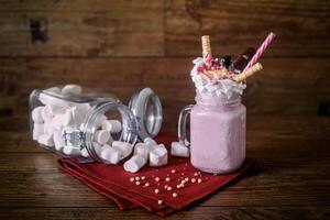 Glass jar of homemade raspberry smoothie cocktail, served with whipped cream, caramel jelly beans and wafer rolls over old dark wood background with jar of marshmallow over background. With copy space photo