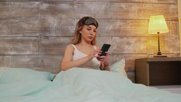 Beautiful woman in pajamas with sleep mask using her phone before going to sleep. video