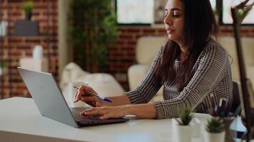 Remote employee at desk doing multitasking, transcribing data from laptop to notepad. Indian woman in apartment trying to complete job tasks before deadline, writing info on paper using pen video