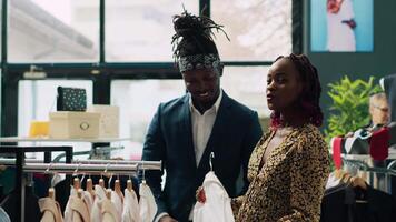 Employees team assisting pregnant woman to find trendy clothes, showing multiple models and colors on hangers. Retail assistants making suggestions for african american customer at store. Camera A. video