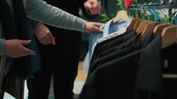 Senior mulher procurando roupas Itens para dela marido, olhando para elegante ternos em cabides às departamento loja. idosos casal compras para a Novo moda coleção às showroom. Câmera b. video