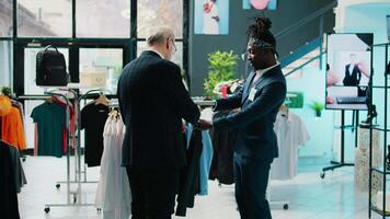 African american store assistant helping regular client with a suit in shopping center, senior customer looking to buy formal clothes. Elderly man shopping for new collection at the mall. Camera B. video