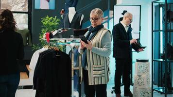 anziano donna controllo sito web azione per trova elementi nel memorizzare a centro commerciale, shopping per il nuovo progettista collezione. cliente utilizzando smartphone App e guardare per formale capi di abbigliamento linea. telecamera b. video