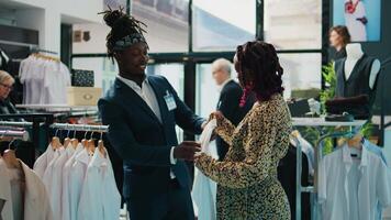 Retail assistant showing new clothing collection to customer, recommending colors and sizes for pregnant woman in department store. African american client asking worker for assistance. Camera B. video