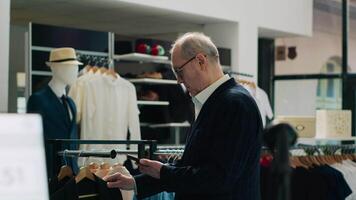 modisch Senior Mann Einkaufen zum Neu Mode Sammlung beim Verkauf speichern, Besuch modern Kleidung Geschäft zu prüfen formal oder beiläufig Artikel. Reich Kunde Verwendet Telefon zu aussehen zum Produkte durch Code. Kamera a. video