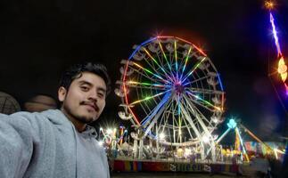 Man taking selfie at Mexican fair with ferris wheel and colorful lights at night photo