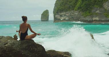 sérénité au milieu de océans rugir. une femme médite sur une rocheux plage, découverte tranquillité près turquoise océan, vagues s'écraser sur falaises. video