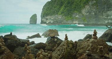 kunstzinnig gestapeld Cairns staan net zo stil schildwachten voordat de rusteloos oceaan golven Aan nusa penida. robuust kustlijn van Bali. sereen zeegezicht. video