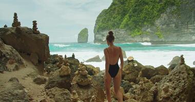 mujer en traje de baño camina descalzo en rocoso playa. contemplando el del océano infinidad. video