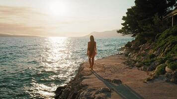 mulher caminhando de a mar às pôr do sol. uma turista passeando ao longo a costeiro caminho com a Sol refletindo fora a adriático mar. Makarska riviera, Croácia. video