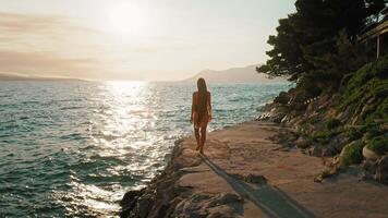 kvinna gående förbi de hav på solnedgång. en turist promenader längs de kust väg med de Sol reflekterande av de adriatisk hav. makarska rivieran, kroatien. video