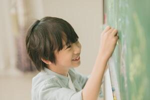 asiático chico contento sonrisa disfrutar educación a colegio en salón de clases escritura responder a pizarrones foto