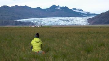 A woman sits in the grass and looks at the glacier and mountains of Iceland. Freedom and travel concept. 4K video