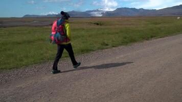 weiblich Tourist mit Rucksack geht durch Schmutz Straße auf Hintergrund von Berge und Gletscher im Island. Freiheit und Reise Konzept. 4k video