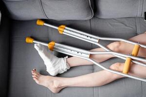 Woman with beautiful, long legs and a cast on her right leg with crutches lies on the sofa, top view photo