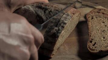 Cutting slice of homemade Crusty rye bread with a sharp knife on wooden board, closeup slow motion video