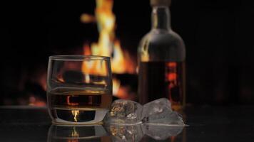 Glass and bottle of whiskey, scotch or bourbon stands on a table against background of a fireplace with a flame. Ice falls into a glass of whiskey. Alcoholic drink and home comfort concept. video