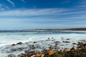 Oceano playa con grande piedras y arena debajo un azul cielo y nubes foto