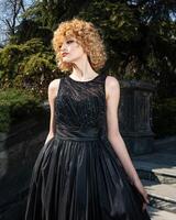 A woman in a black dress stands on steps in front of trees. photo