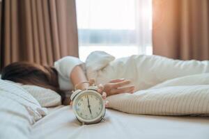 alarm clock and asian woman hand stop time in bed while sleeping, young adult female wake up late in the early morning. Relaxing , sleepy, daily routine and have a nice day concepts photo